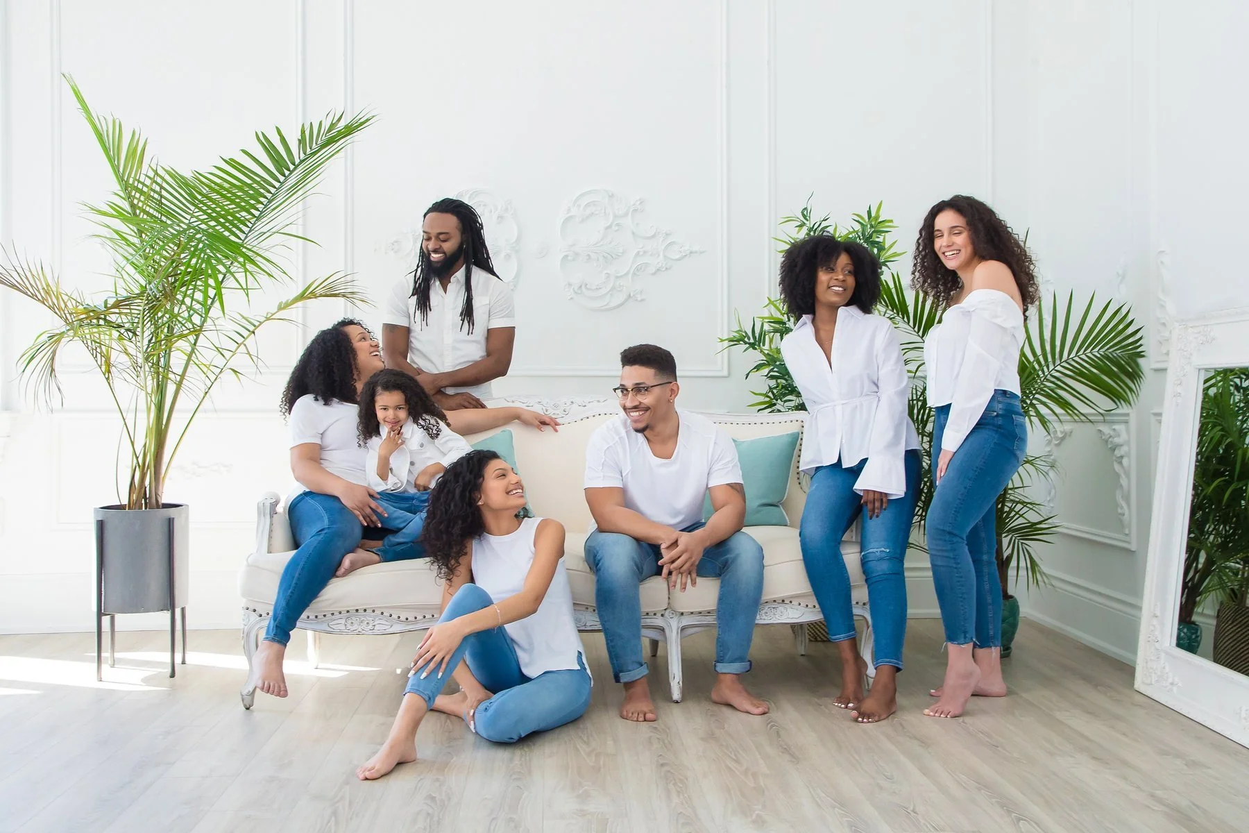 Group of adults and children in white tops and jeans laughing and talking in a bright, plant-filled living room with a white sofa.