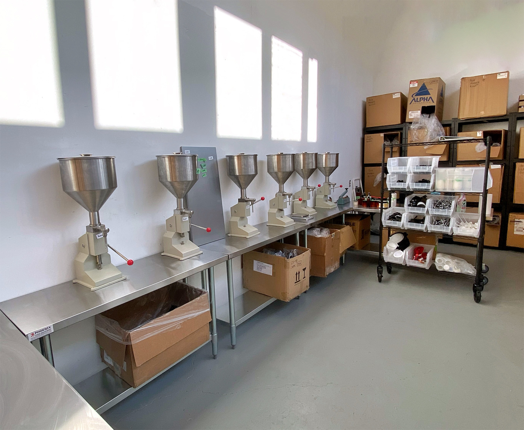 Row of stainless steel hopper grinders lined up on a stainless counter in a storage/workroom with shelves and labeled boxes in the background.