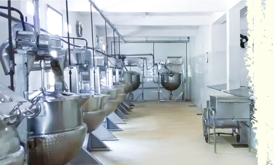 Row of large stainless steel industrial mixing vats and equipment in a clean food processing facility corridor, with pipes overhead and white walls far end of room.